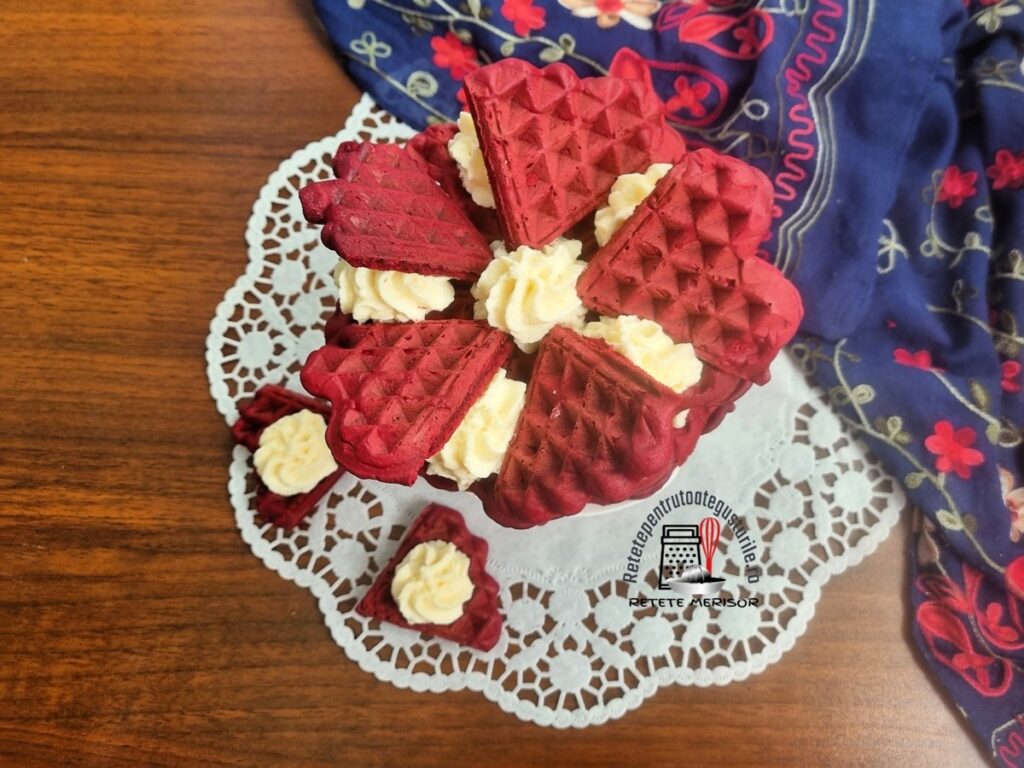 Tortuleț din vafe red velvet cu cremă de cocos pe un platou alb cu picior, alături de două vafe inimioară cu cremă