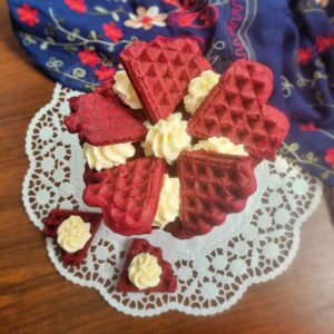 Tortuleț din vafe red velvet cu cremă de cocos pe un platou alb cu picior, alături de două vafe inimioară cu cremă