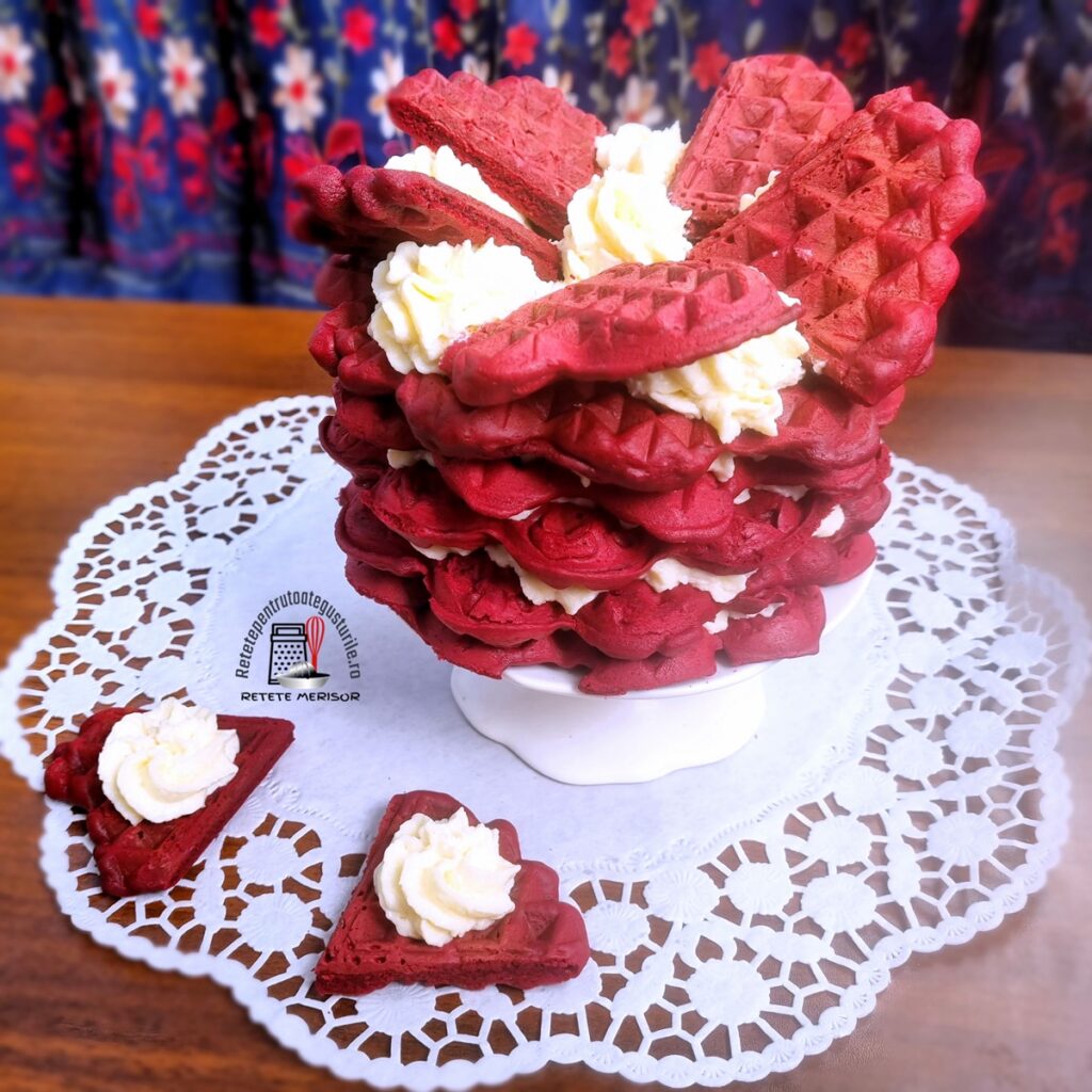 Tortuleț din vafe red velvet cu cremă de cocos pe un platou alb cu picior, alături de două vafe inimioară cu cremă